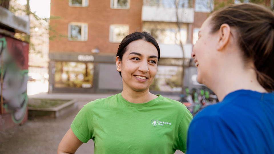 En kvinna i grön t-shirt och en kvinna i blå t-shirt kollar på varandra och samtalar utanför ett bostadshus.