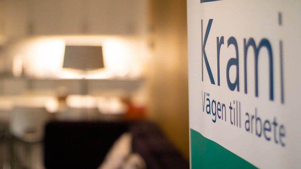 En skylt med texten Krami Vägen till arbete. I bakgrunden syns ett rum med soffa och lampa.